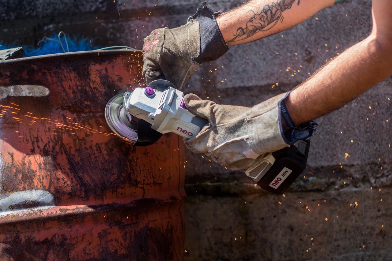 Worker uses angle grinder to cut metal drum, safety gloves visible.